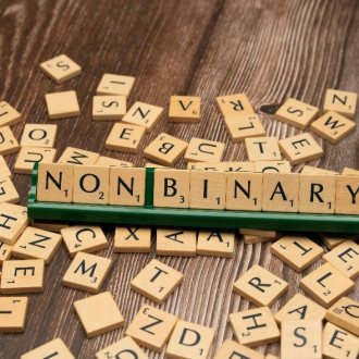 Auf einer Holzfläche liegen kleine Scrabble – Spielebuchstaben. Auf einer Ablage ist das Wort NON BINARY (Nicht binär) aneinandergereiht.